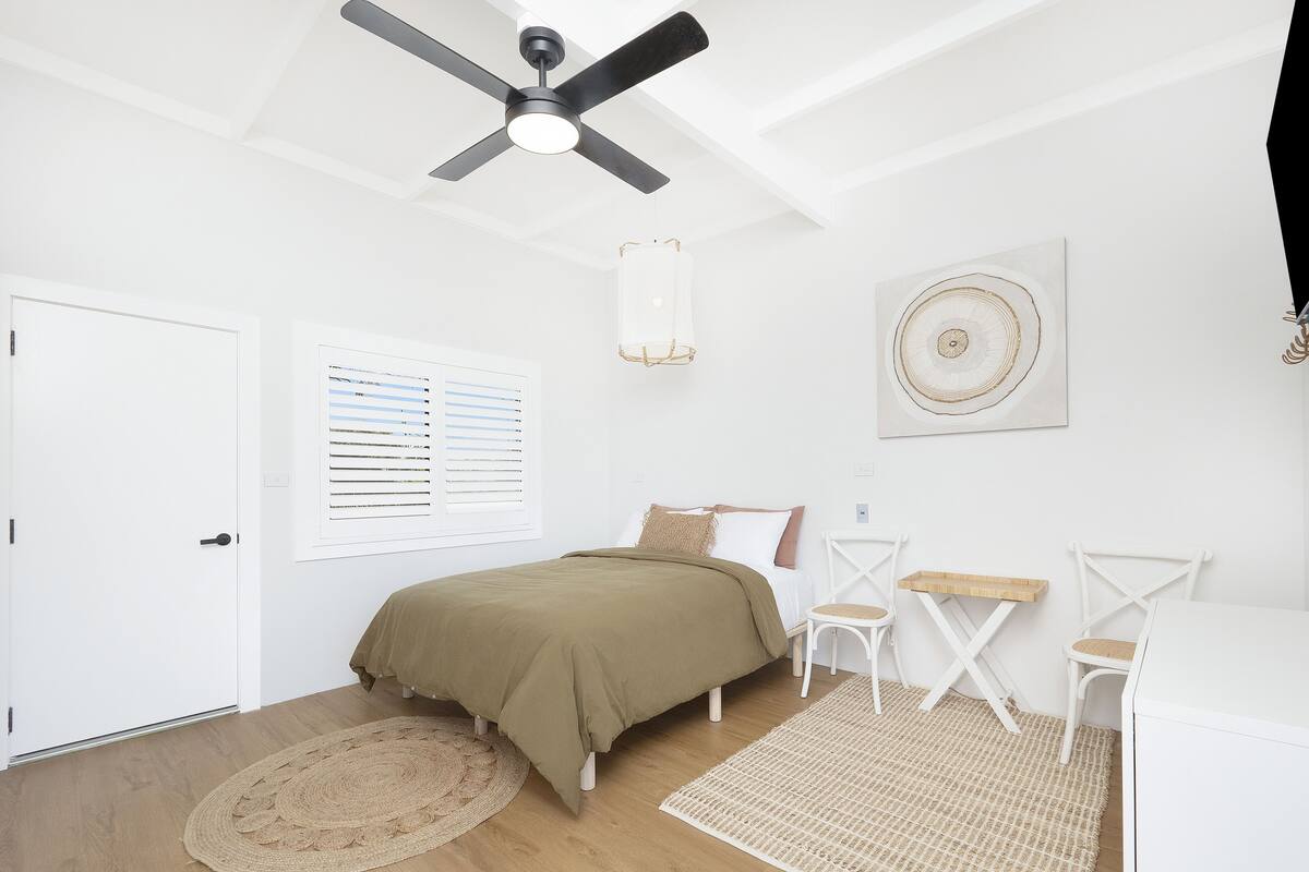 A bedroom with a bed, a ceiling fan, a window with blinds, a wall-mounted artwork, and a small table with chairs.