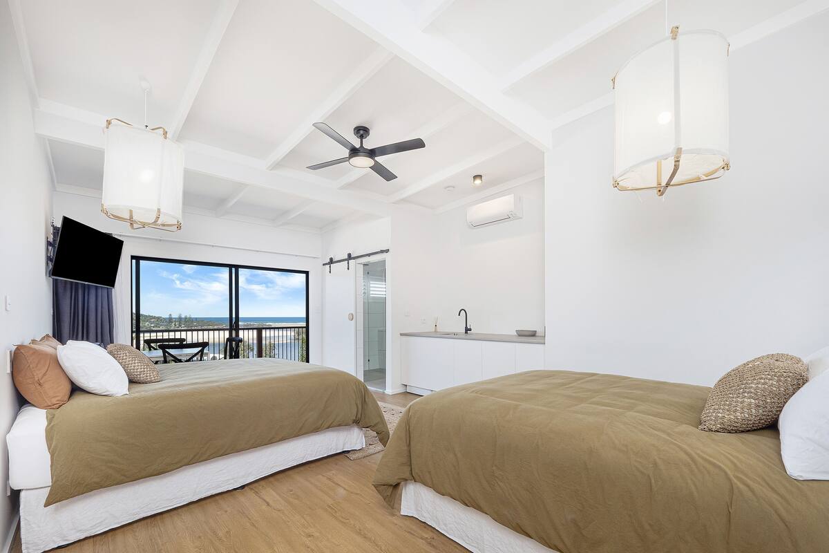 A modern hotel room with two beds, a balcony with a view, a ceiling fan, and a kitchen area.