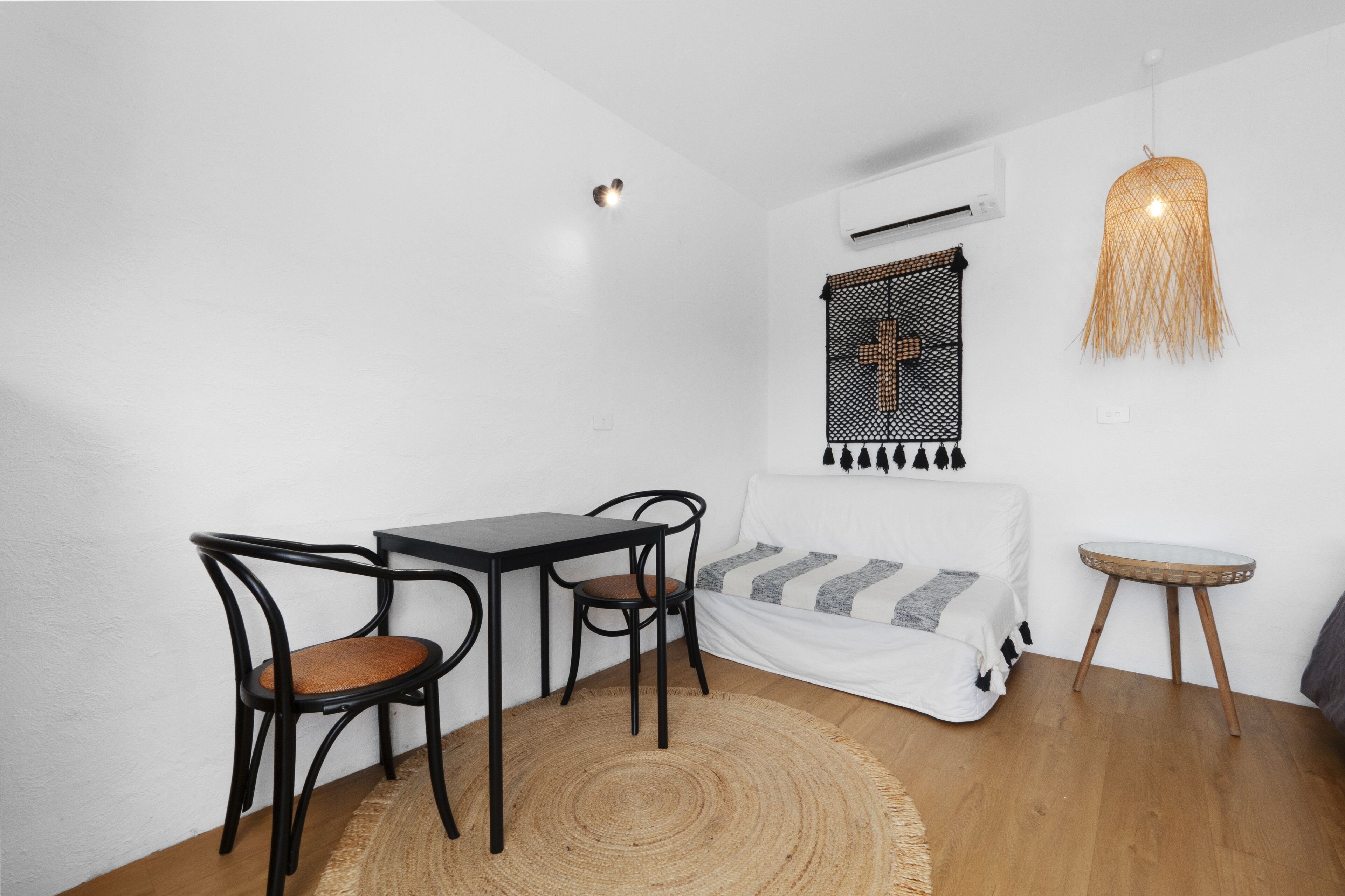 Una habitación pequeña y bien iluminada con una cama, una mesa de comedor con sillas, un tapete tejido y una lámpara colgante.