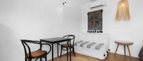 Una habitación pequeña y bien iluminada con una cama, una mesa de comedor con sillas, un tapete tejido y una lámpara colgante.