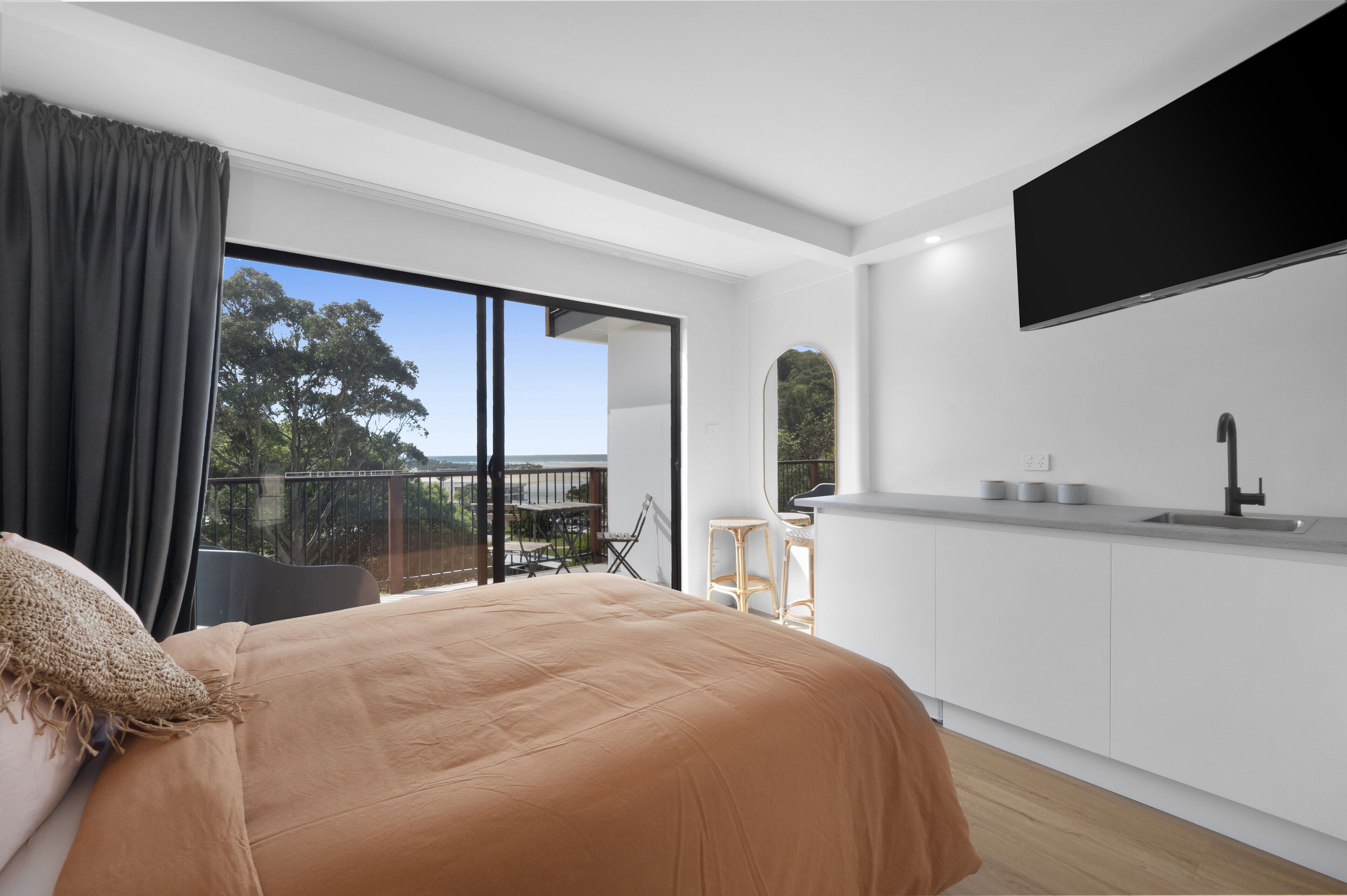 A modern bedroom with a large bed, a balcony with a view, a kitchen area with a flat-screen TV, and a dining area.