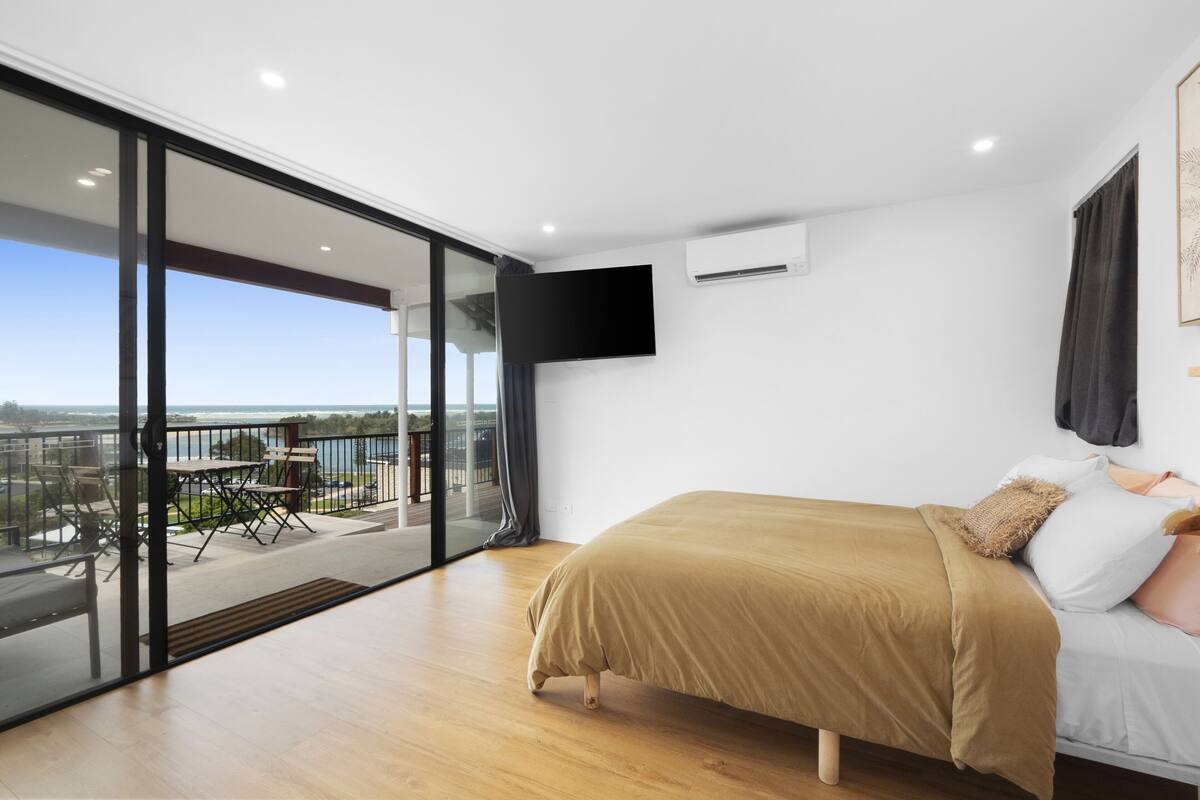 A modern bedroom with a large bed, a TV mounted on the wall, and a balcony with a view of the sea.