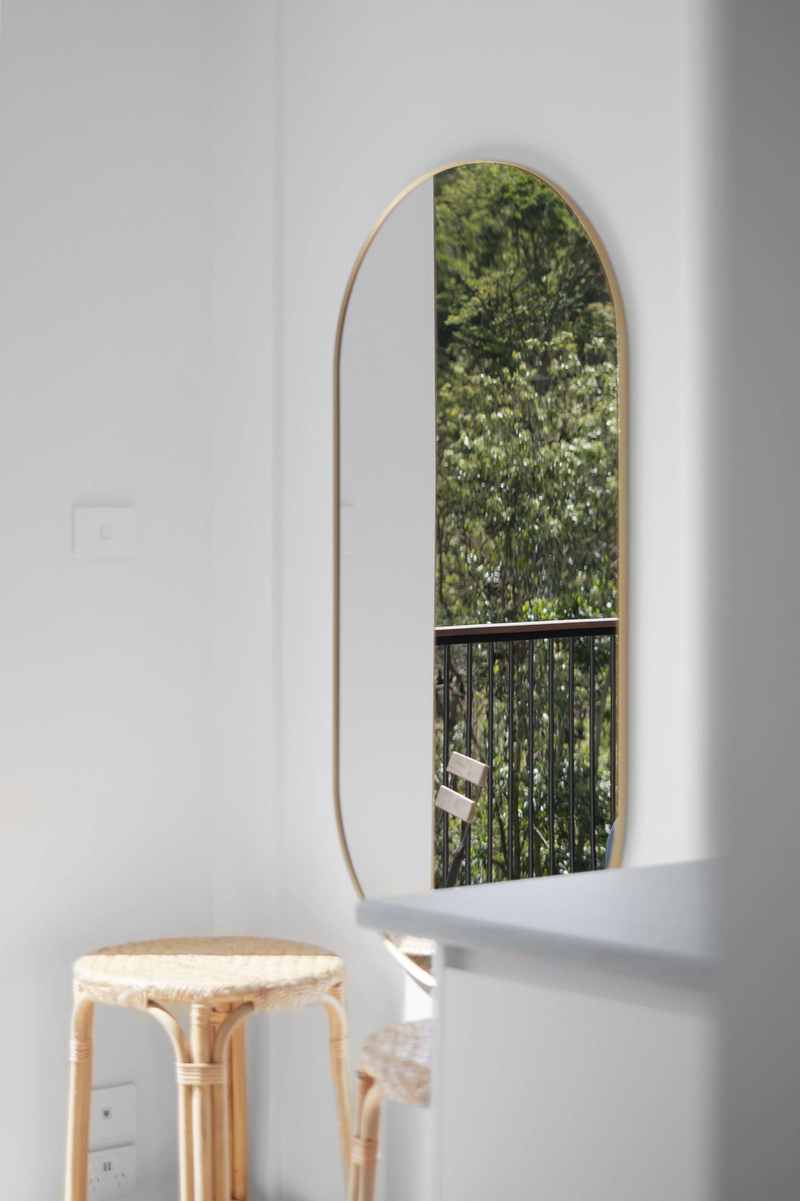 A room with a white wall, a wicker chair, and a mirror reflecting a balcony with greenery.