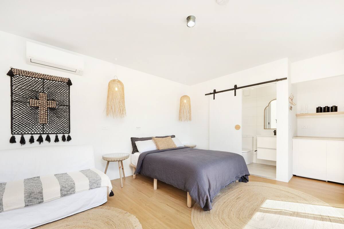 A modern bedroom with a bed, a small round table, a sofa, and a bathroom visible through an open door.