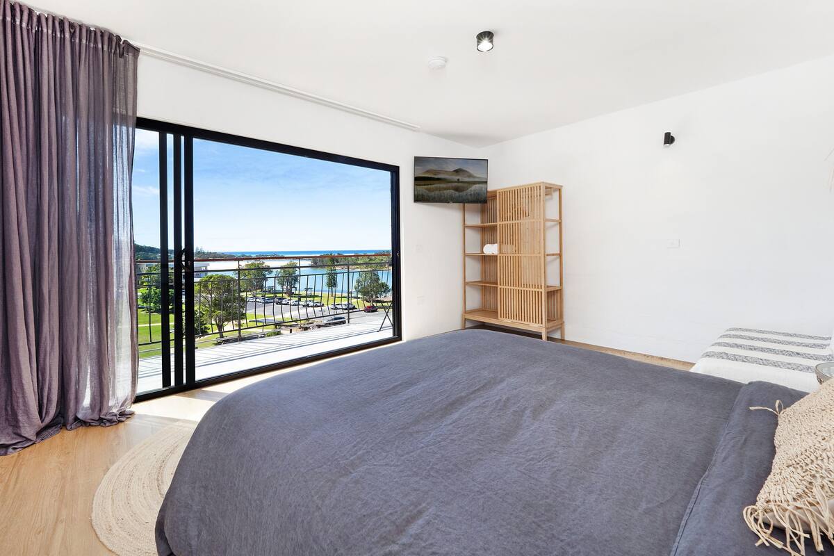 A bedroom with a large bed, a wooden bookshelf, a TV on the wall, and a view of a body of water and buildings.