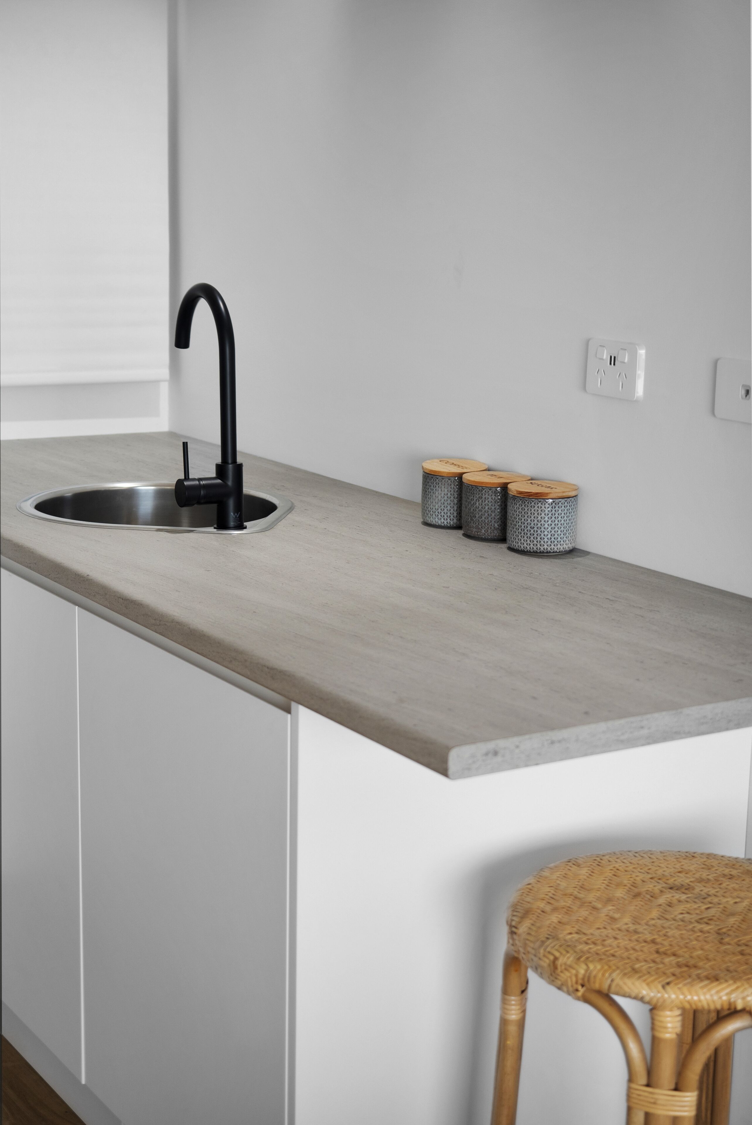 Une cuisine dotée d’un robinet noir moderne, d’un plan de travail gris clair, de trois récipients décorés et d’un tabouret en osier.