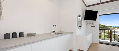 Cocina moderna con armarios blancos, encimera gris, grifo negro y un lavamanos pequeño. Hay tres recipientes negros sobre la encimera. Un televisor de pantalla plana está montado en la pared sobre el lavamanos.