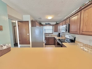 A compact kitchen with wooden cabinets, a stainless steel refrigerator, a microwave, and a stove. - Things are different at the beach ~~~ 🏖️ Come enjoy the breeze! (Gulf Shores)