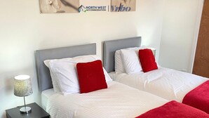 A hotel room with two beds, each with white bedding and red pillows, a bedside lamp, and a wall-mounted sign for North West Accommodation. - 1 Bedroom St Helens Apartment | Free WiFi | Fully Furnished (St Helens)