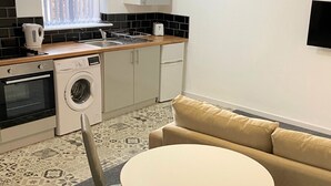 A compact kitchen with a washing machine, a toaster, and a sink. There's a beige sofa and a small round table. - 1 Bedroom St Helens Apartment | Free WiFi | Fully Furnished (St Helens)