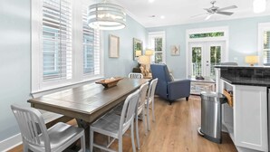 A modern kitchen with a dining area, featuring a wooden table, white chairs, a blue armchair, and a ceiling fan. - Spacious 30A Home Steps to Beach With King Suites and Loft Space (Panama City Beach)