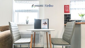 A modern room with two grey cushioned chairs, a small white table, and a window with blinds. A welcome sign and a fire blanket are visible on the wall.