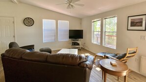 Ein Wohnzimmer mit einem braunen Sofa, einem Fernseher auf einem Schrank, einem Deckenventilator und zwei Fenstern mit Jalousien.