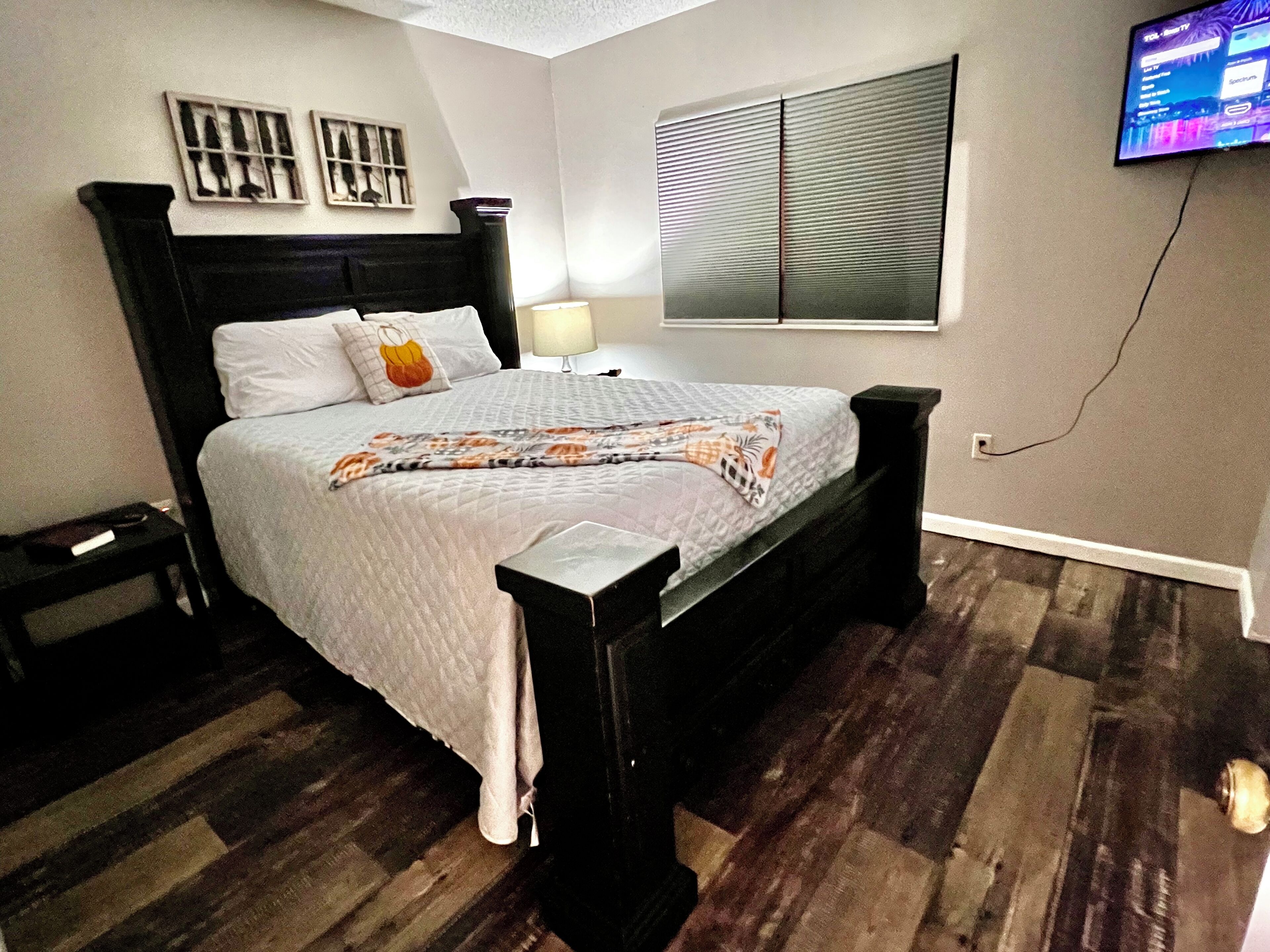 Un dormitorio ordenado con cama, mesitas de noche, televisor montado en la pared y ventana con persianas.
