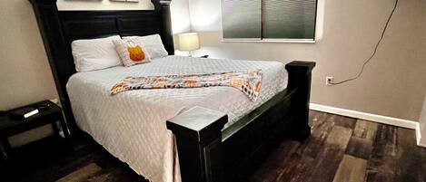 Un dormitorio ordenado con cama, mesitas de noche, televisor montado en la pared y ventana con persianas.