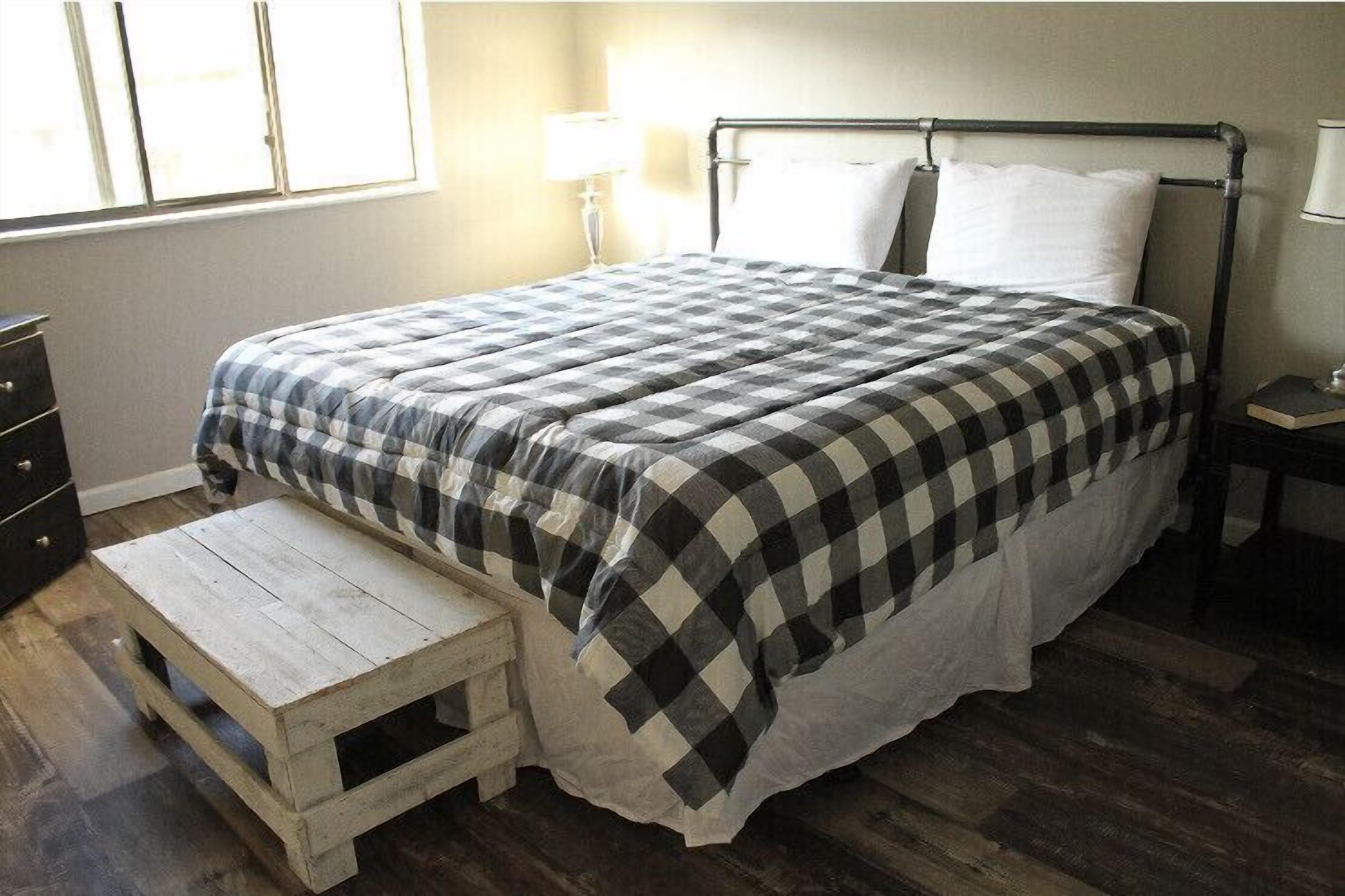 Een net opgemaakt bed met een geruit sprei, een nachtkastje met een lamp en een klein houten krukje.