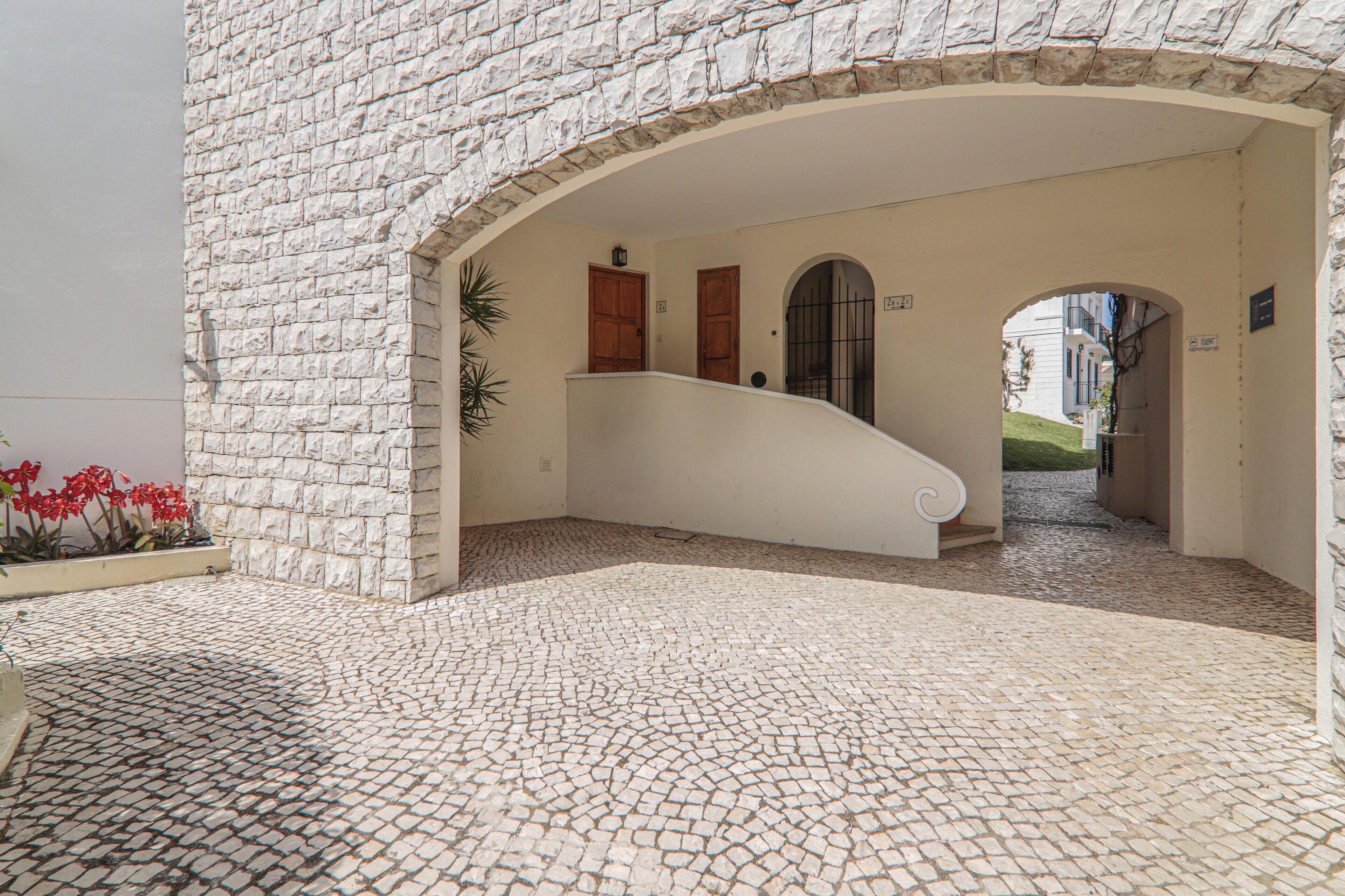 Une entrée en arc surmontée d’un sol pavé, d’un mur blanc et d’une porte portant un numéro.