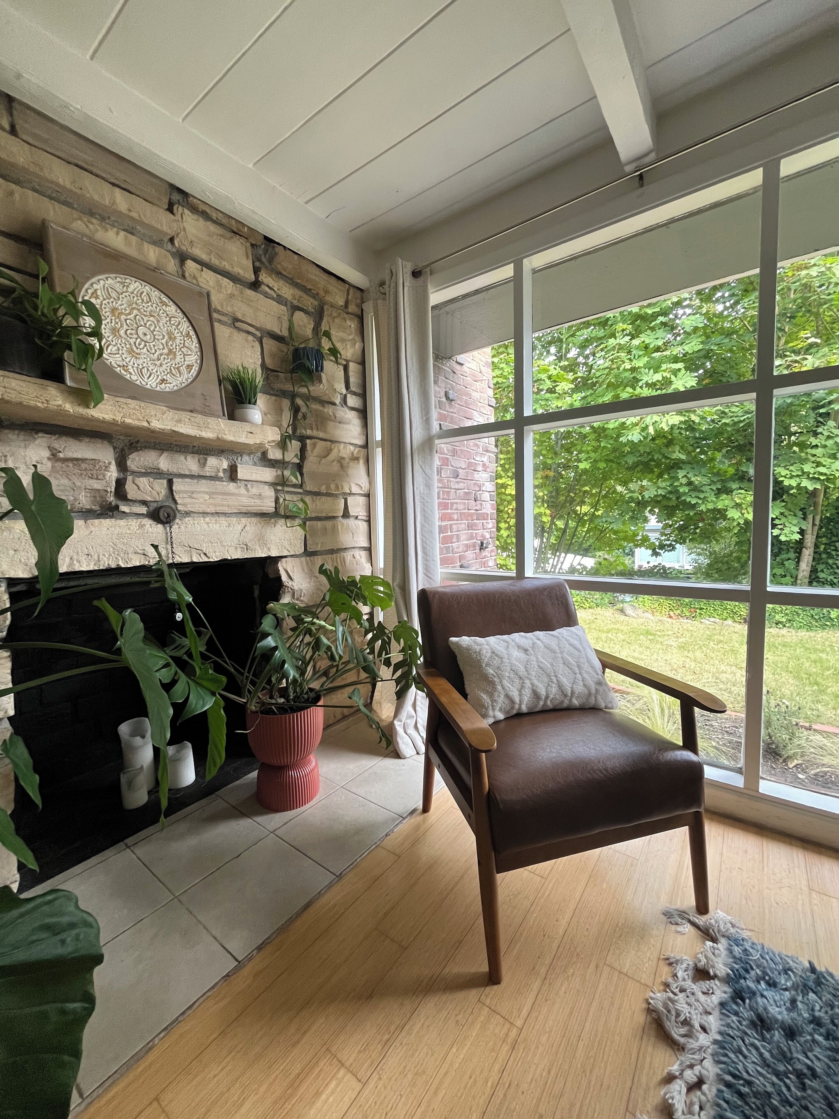 A cozy room with a stone fireplace, a leather armchair, and a large window offering a view of greenery.