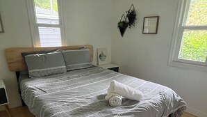A modern bedroom with a wooden bed frame, grey bedding, and a neatly made white towel. There are two bedside tables with lamps, a window with blinds, and a wall-mounted plant. - Cool Mid-Century Bungalow!  (Vancouver)
