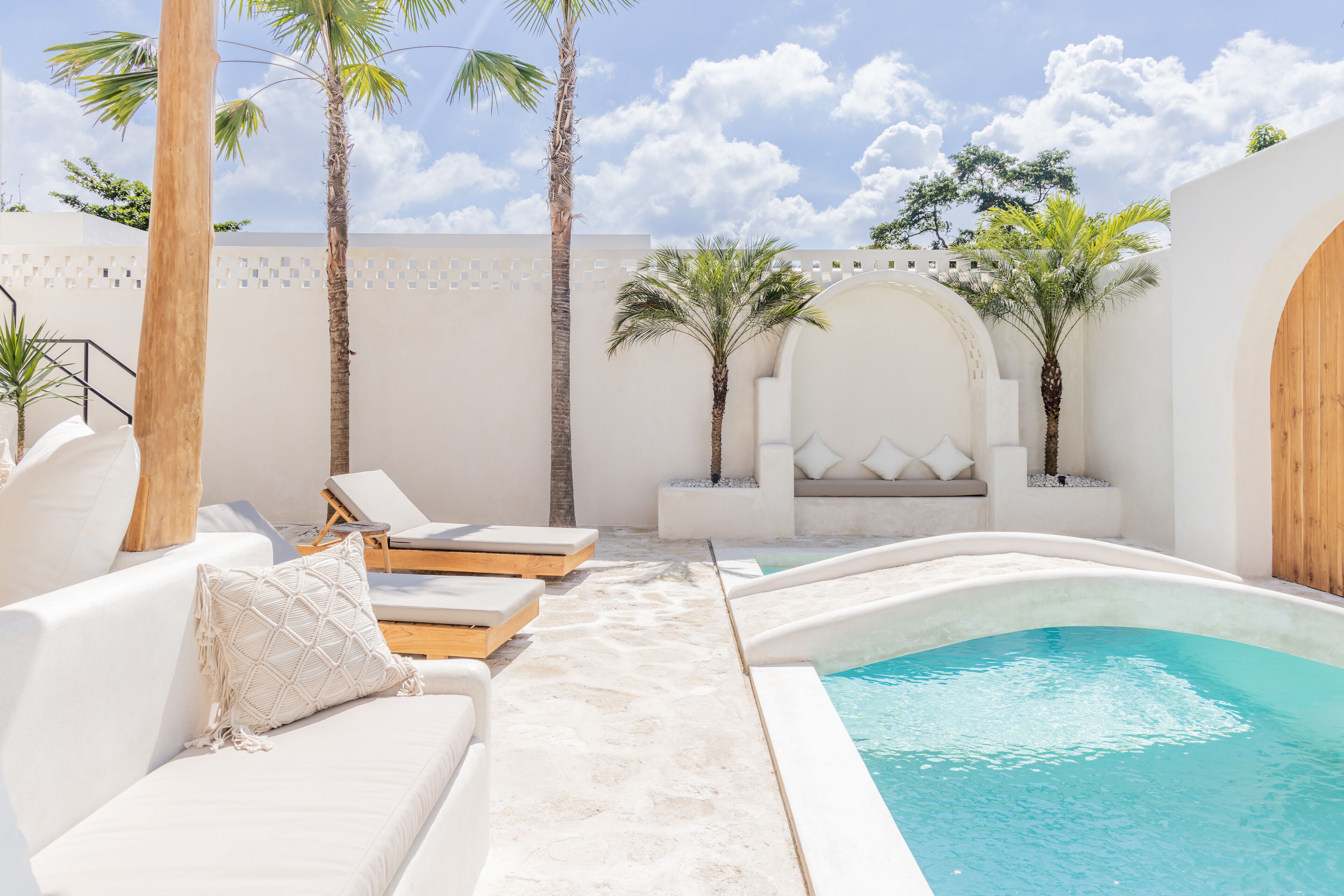 Zona junto a la piscina con sillas reclinables blancas, una piscina pequeña y palmeras.