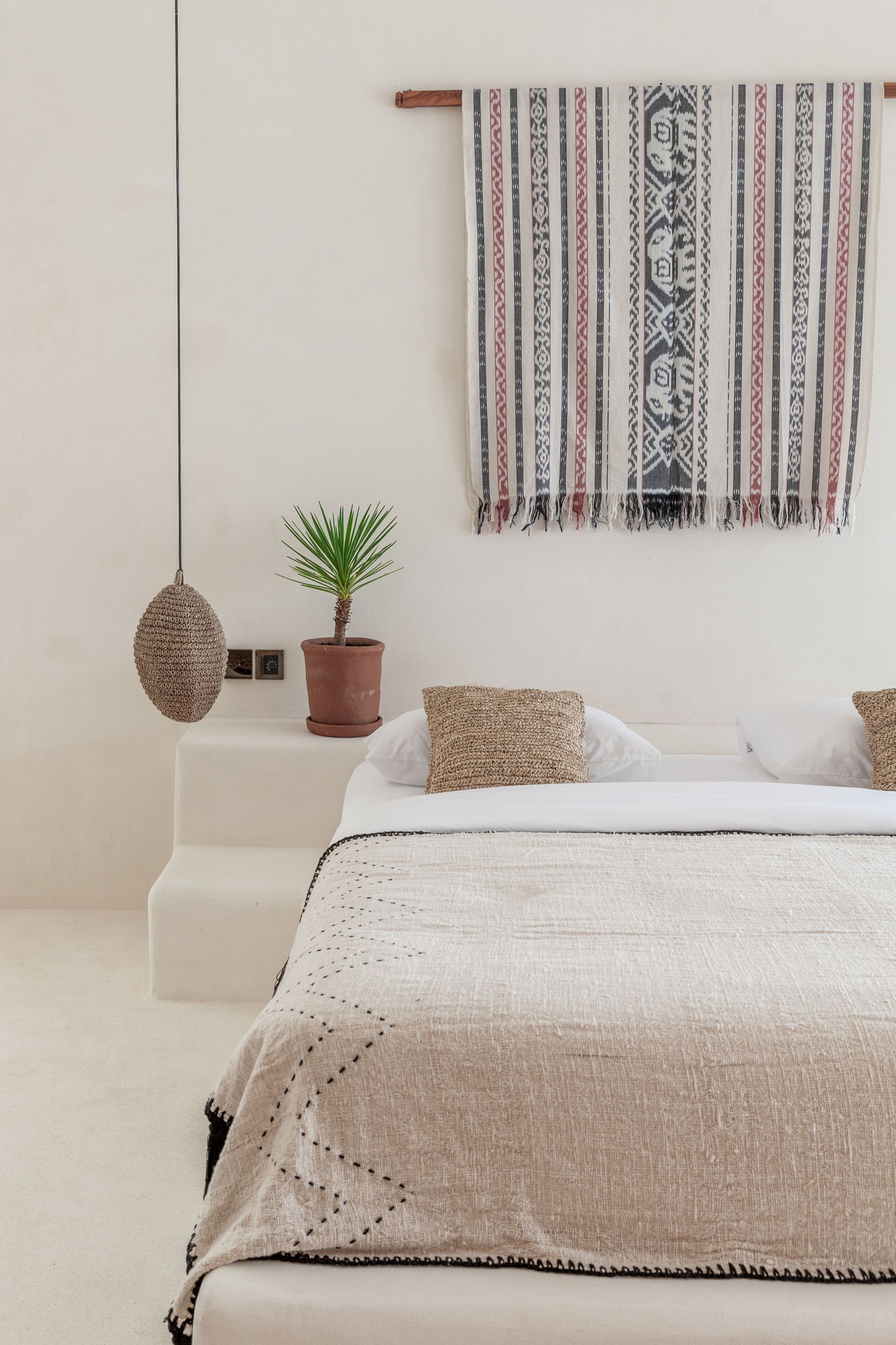 A minimalist bedroom with a bed, a hanging light, a potted plant, and a patterned wall hanging.
