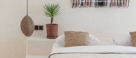 A minimalist bedroom with a bed, a hanging light, a potted plant, and a patterned wall hanging.