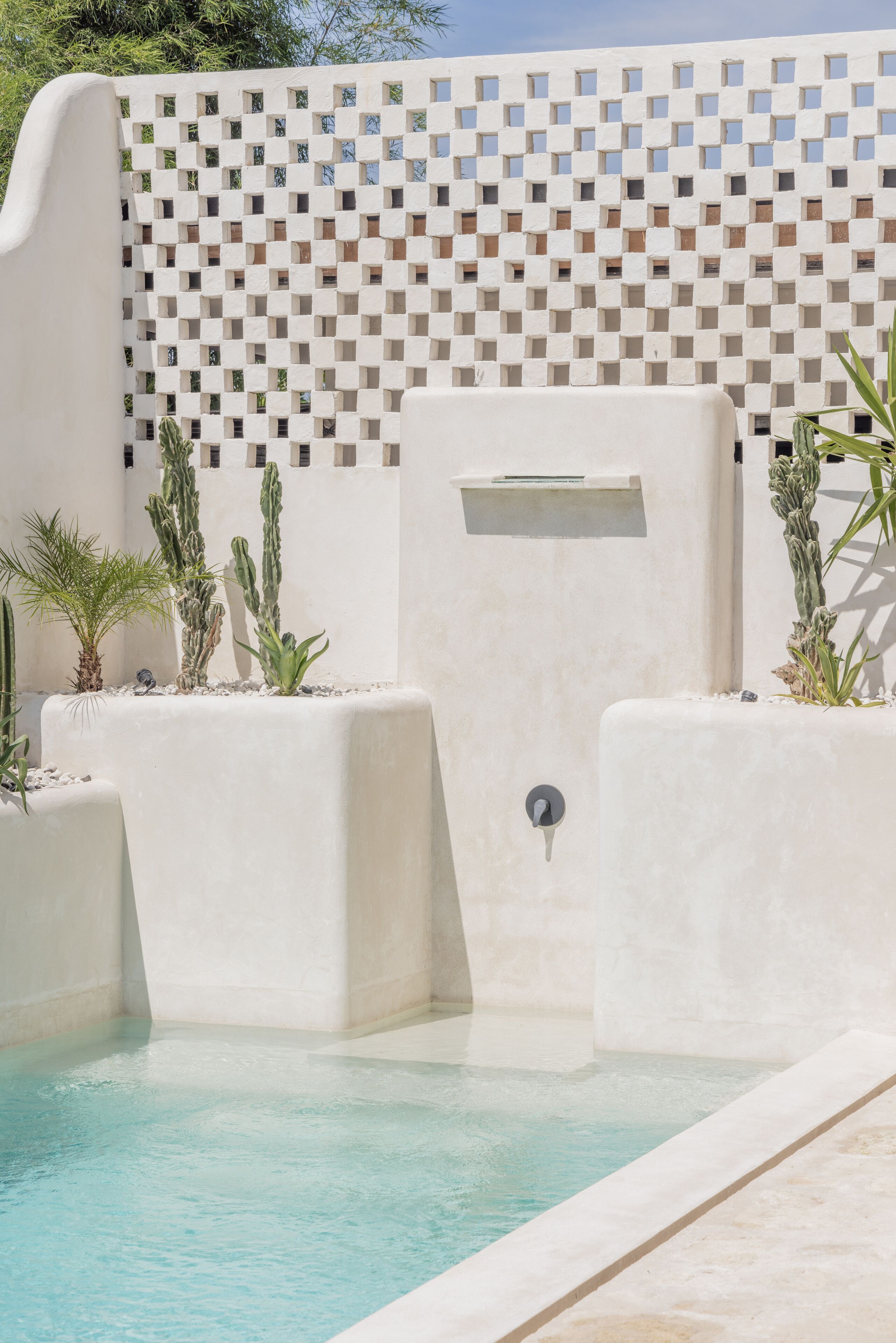 Un área de piscina con suelo de baldosas blancas, pared blanca con diseño geométrico y varios cactus en macetas.