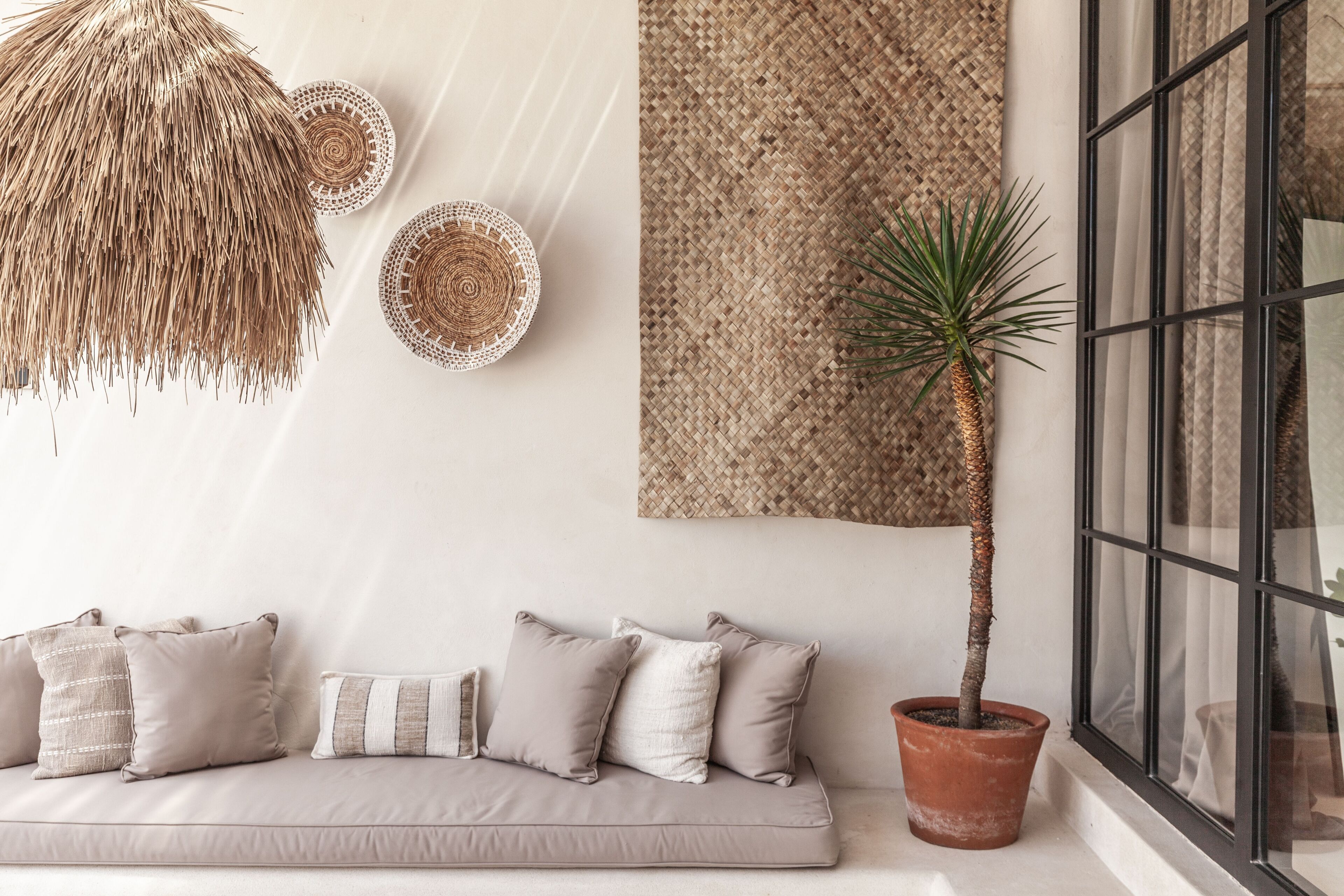A modern living room with a beige sofa, decorative pillows, a woven wall hanging, a potted plant, and a large window with curtains.