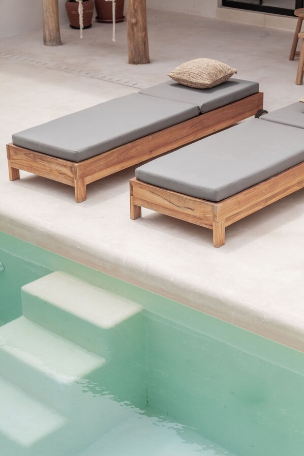 Un área junto a la piscina con dos sillones de madera, cojines grises y una pequeña almohada tejida en uno de ellos.