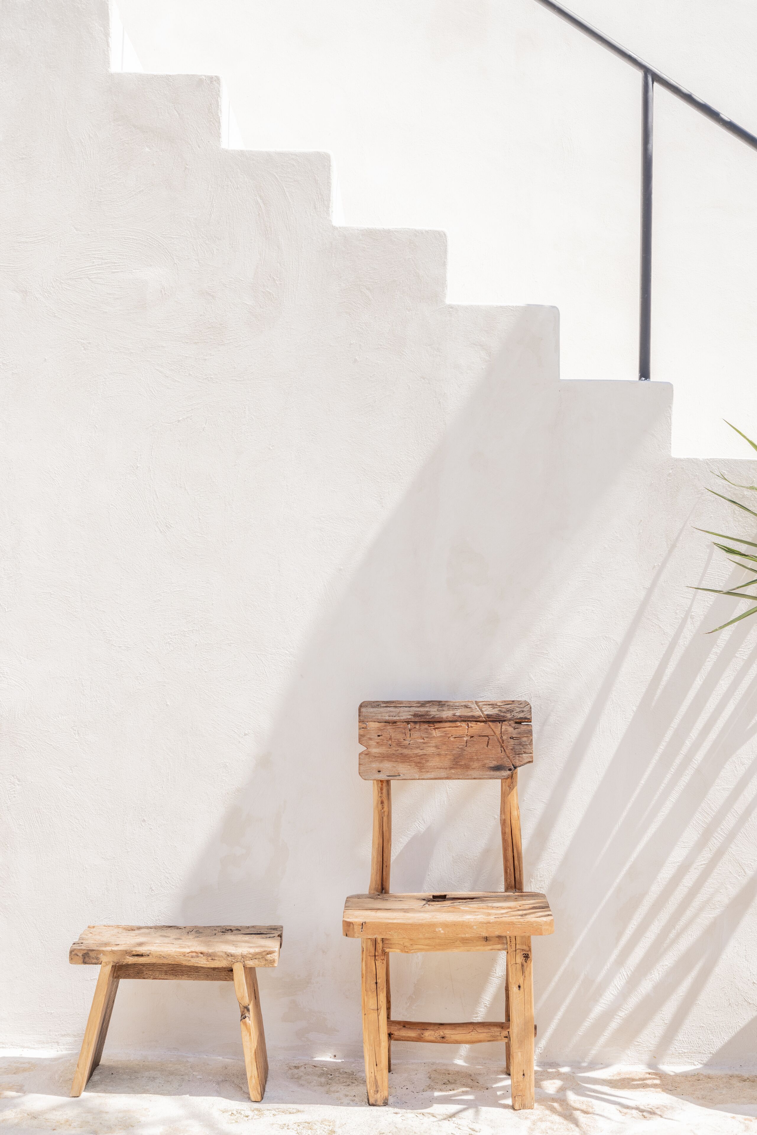 A wooden chair and stool against a white wall with a staircase.