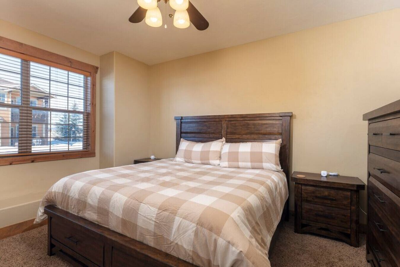 A bedroom with a large bed, wooden headboard, two bedside tables, a dresser, and a window with blinds.