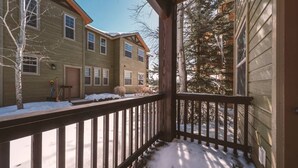 Eine Holzterrasse mit Schnee, ein Haus mit brauner Tür und ein Baum im Hintergrund.