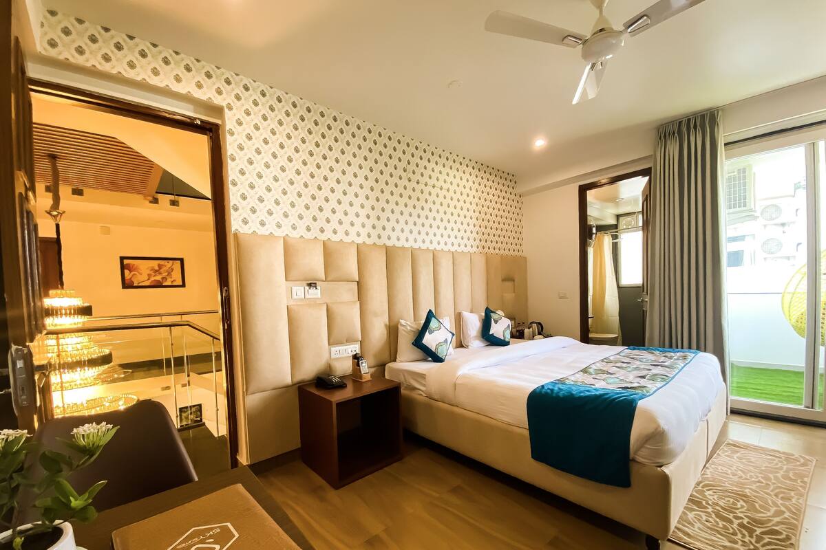 A hotel room with a large bed, a bedside table, a desk with a chair, a potted plant, and a view of the outside.