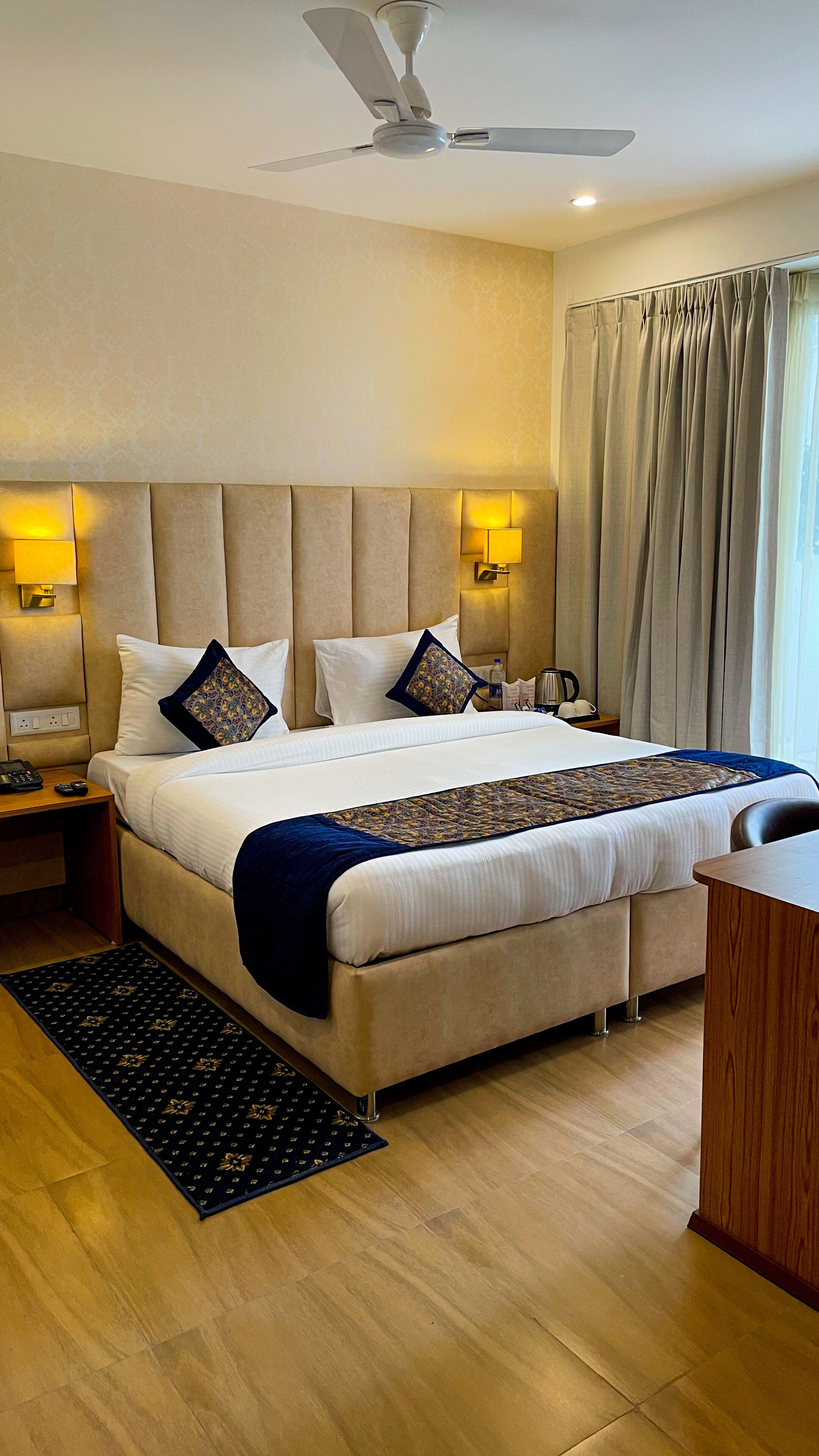 Habitación de hotel con una cama grande, ventilador de techo, lámparas de noche, mesita de noche, escritorio y ventana con cortinas.