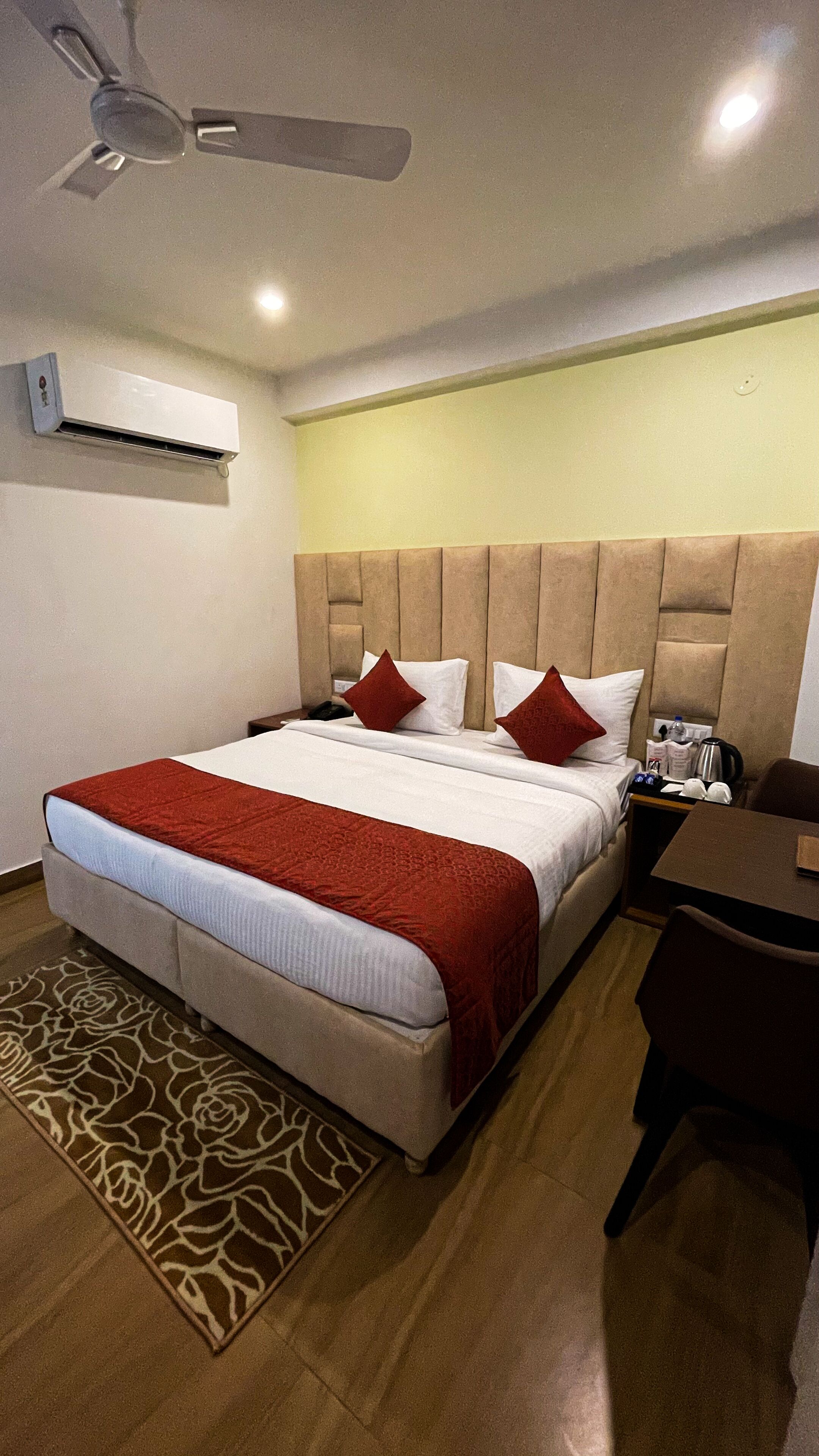 A hotel room with a large bed, a ceiling fan, an air conditioning unit, a bedside table with a lamp, a chair, and a patterned rug.