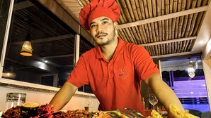 A chef in a red uniform with a chef's hat, standing behind a table with various dishes and a wine glass. - Sky Suites Huda City Center (Gurugram)
