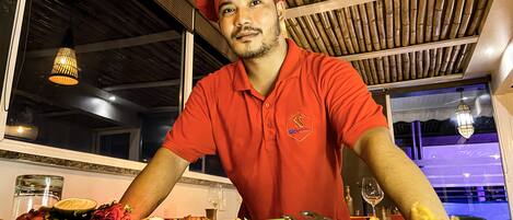 A chef in a red uniform with a chef's hat, standing behind a table with various dishes and a wine glass.