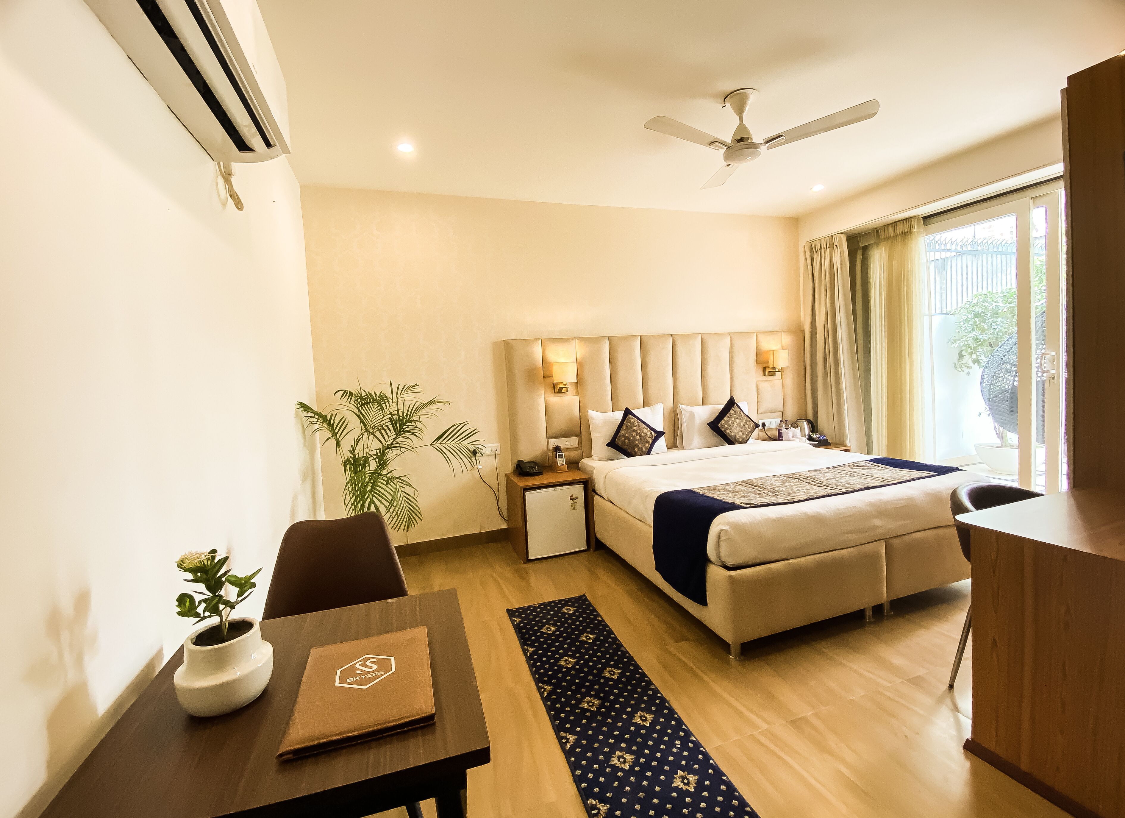 A hotel room with a bed, bedside table, chair, desk, and a potted plant.