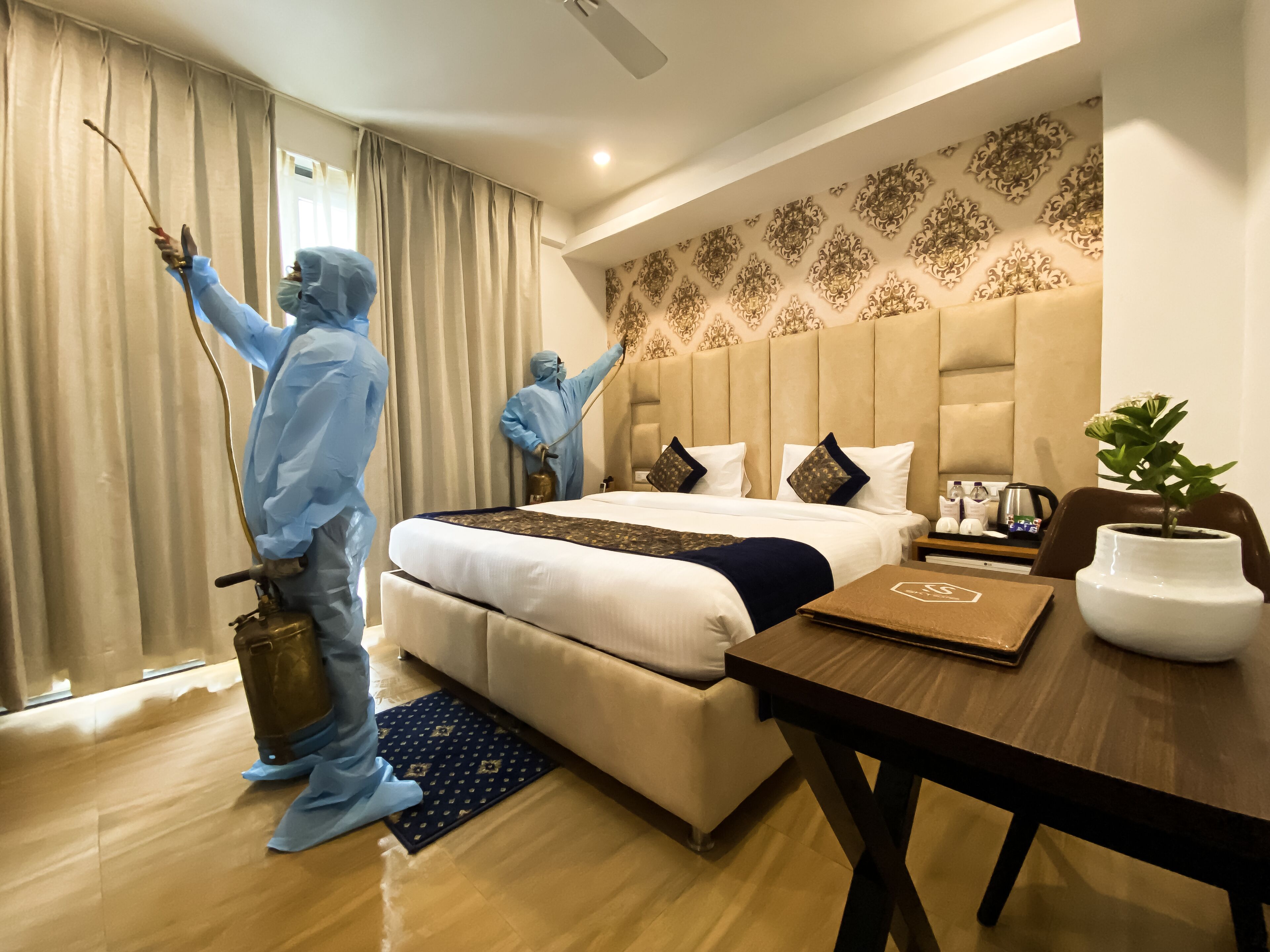 Habitación de hotel con una cama grande, una mesa de madera, una planta en maceta y dos personas con trajes protectores limpiando la habitación.