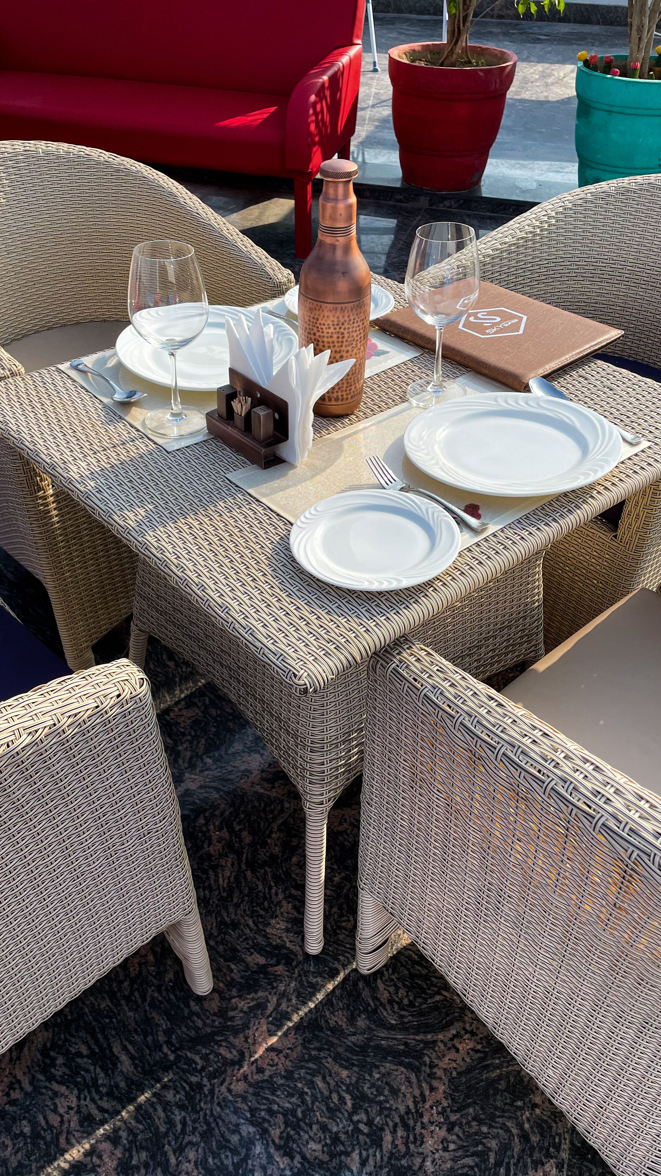 A wicker outdoor dining set with plates, glasses, and a bottle, surrounded by wicker chairs and a red sofa.
