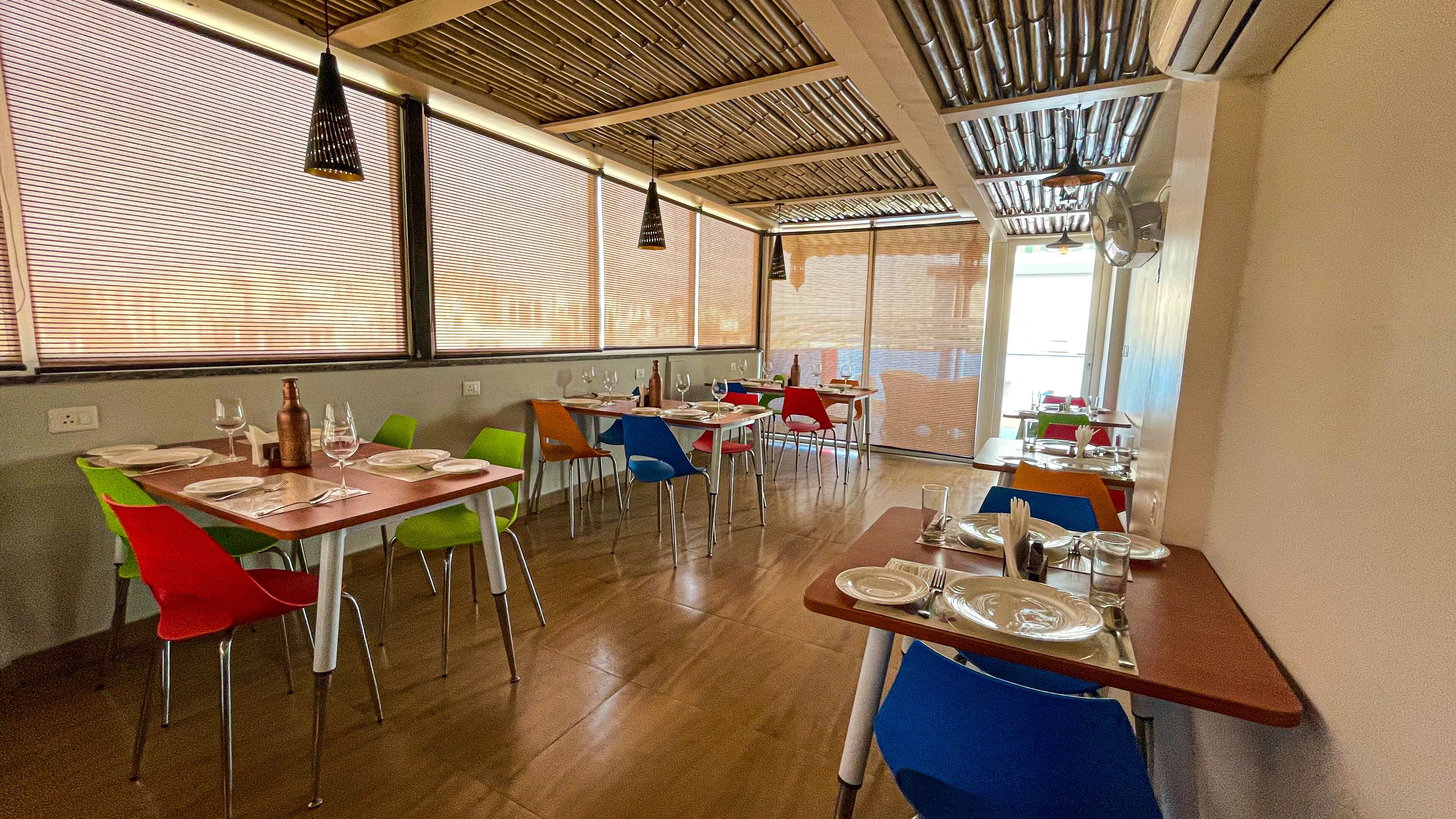 Un restaurante con mesas y sillas de madera, ventanas amplias con persianas y lámparas colgantes.