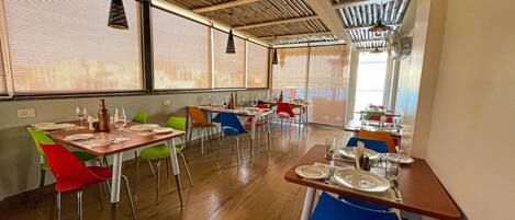Un restaurante con mesas y sillas de madera, ventanas amplias con persianas y lámparas colgantes.