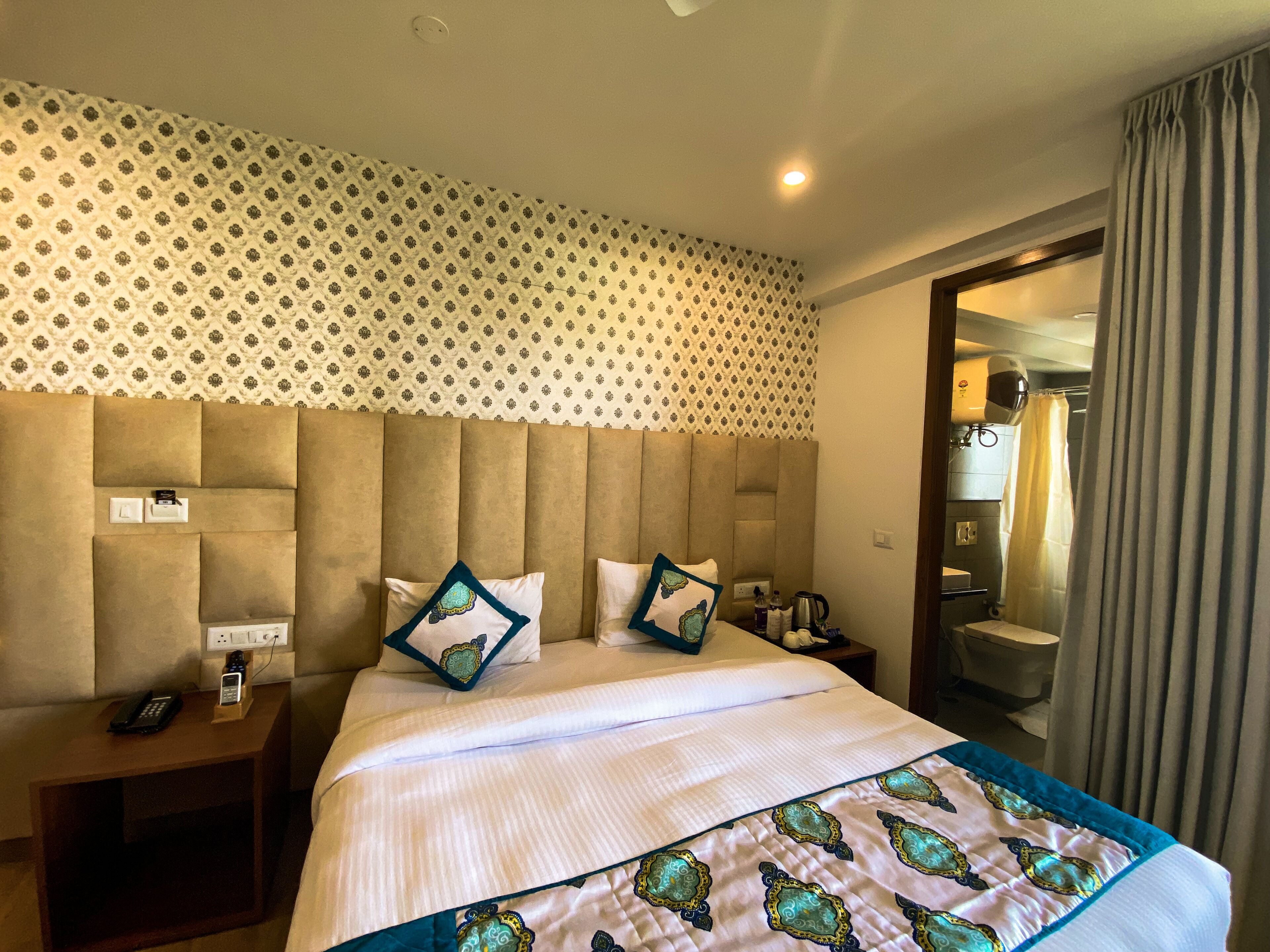 A hotel room with a large bed, a headboard with multiple sections, a bedside table with a phone, and a bathroom visible through an open door.