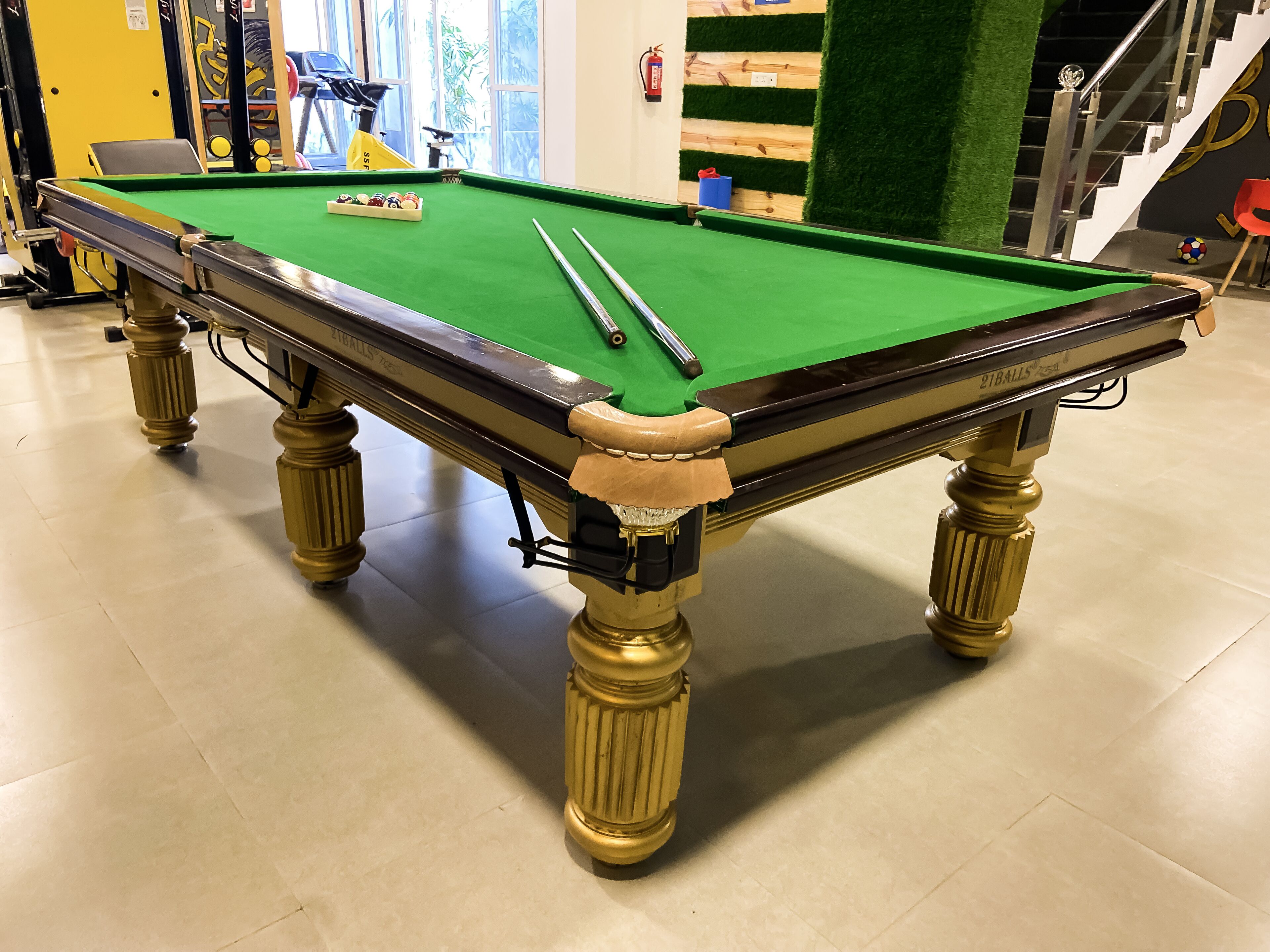 Une table de billard recouverte d’un drap vert, soutenue par des pieds dorés, dans une pièce dont le sol est carrelé et où l’on aperçoit un escalier au fond.