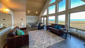 Ein geräumiges Wohnzimmer mit großem Fenster mit Blick auf einen Strand, einer Holzzieldecke und einem modernen Interieur.