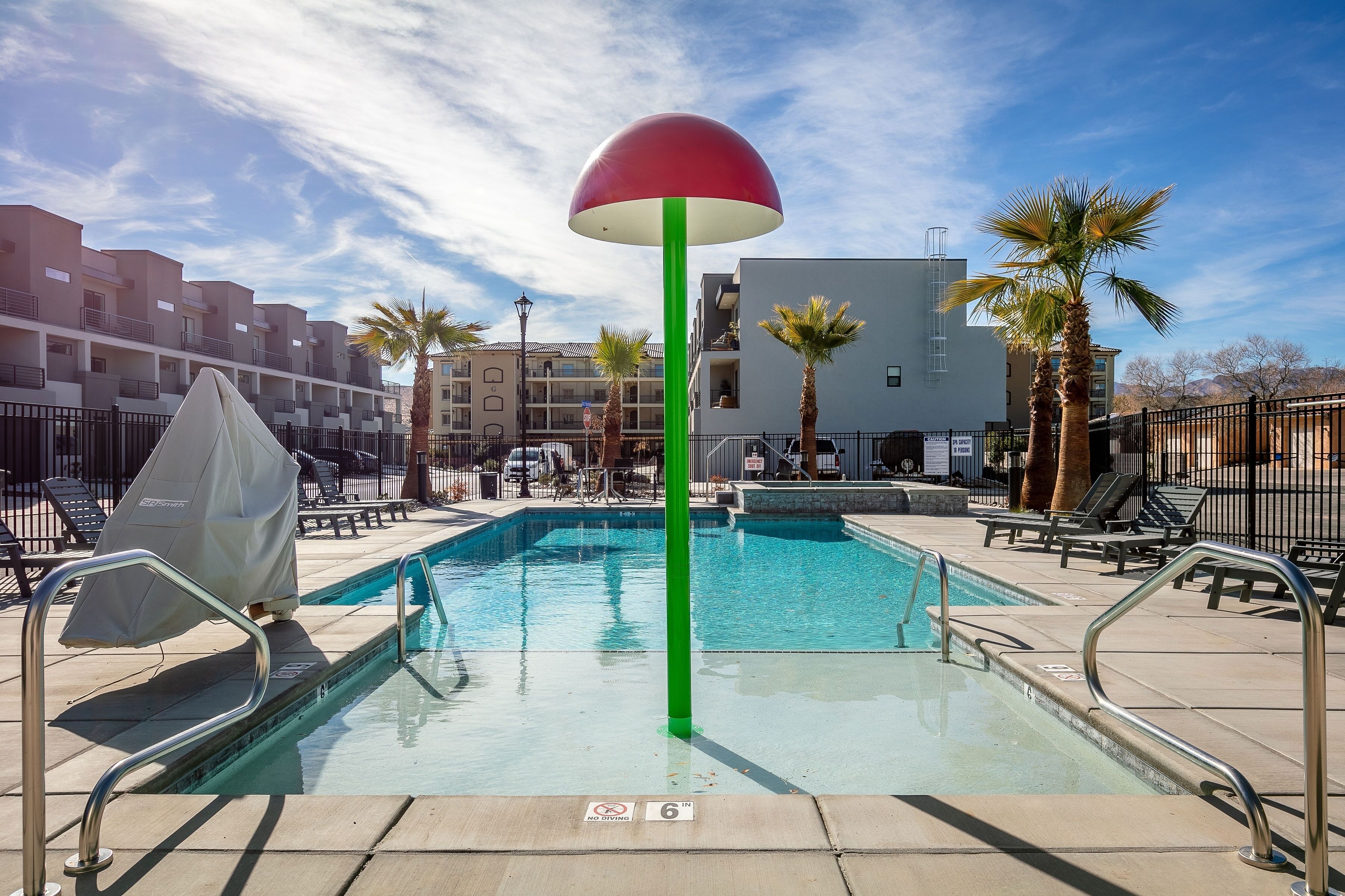 Una piscina con un poste verde y una sombrilla roja, rodeada de sillas de descanso y palmeras.