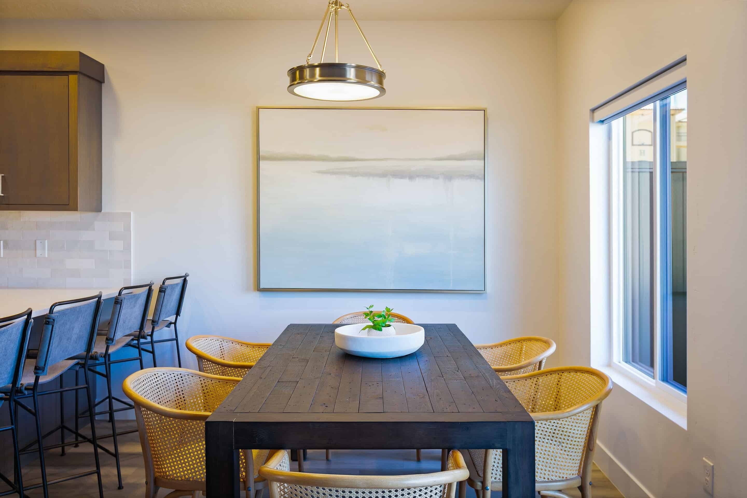 A dining area with a dark wooden table, wicker chairs, a white bowl with a plant, a hanging light fixture, and a large framed picture on the wall.