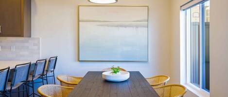 A dining area with a dark wooden table, wicker chairs, a white bowl with a plant, a hanging light fixture, and a large framed picture on the wall.