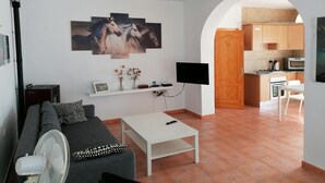 A living room with a sofa, a coffee table, a TV, and a painting of horses on the wall. There is a kitchen visible through an archway.
