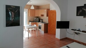 A modern kitchen with wooden cabinets, a microwave, and a stove. There is a dining area with a white table and chairs. A TV is mounted on the wall.